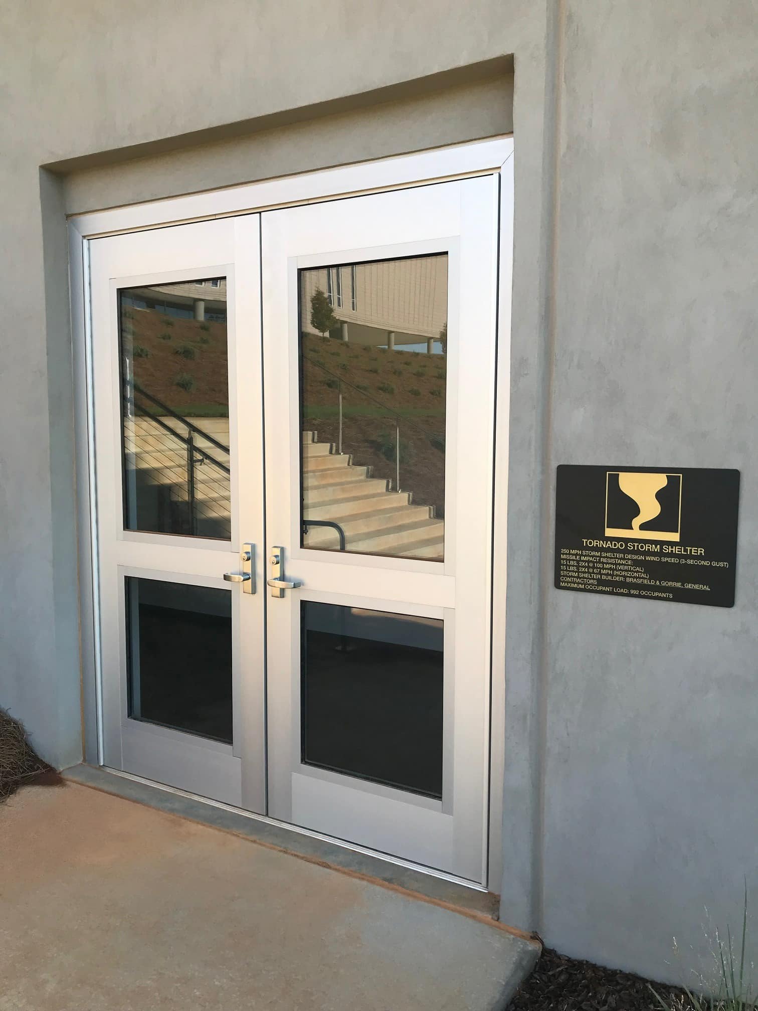 A sturdy, double door with a metal frame leads to a tornado storm shelter. A plaque beside it indicates "Tornado Storm Shelter" with a tornado icon.
