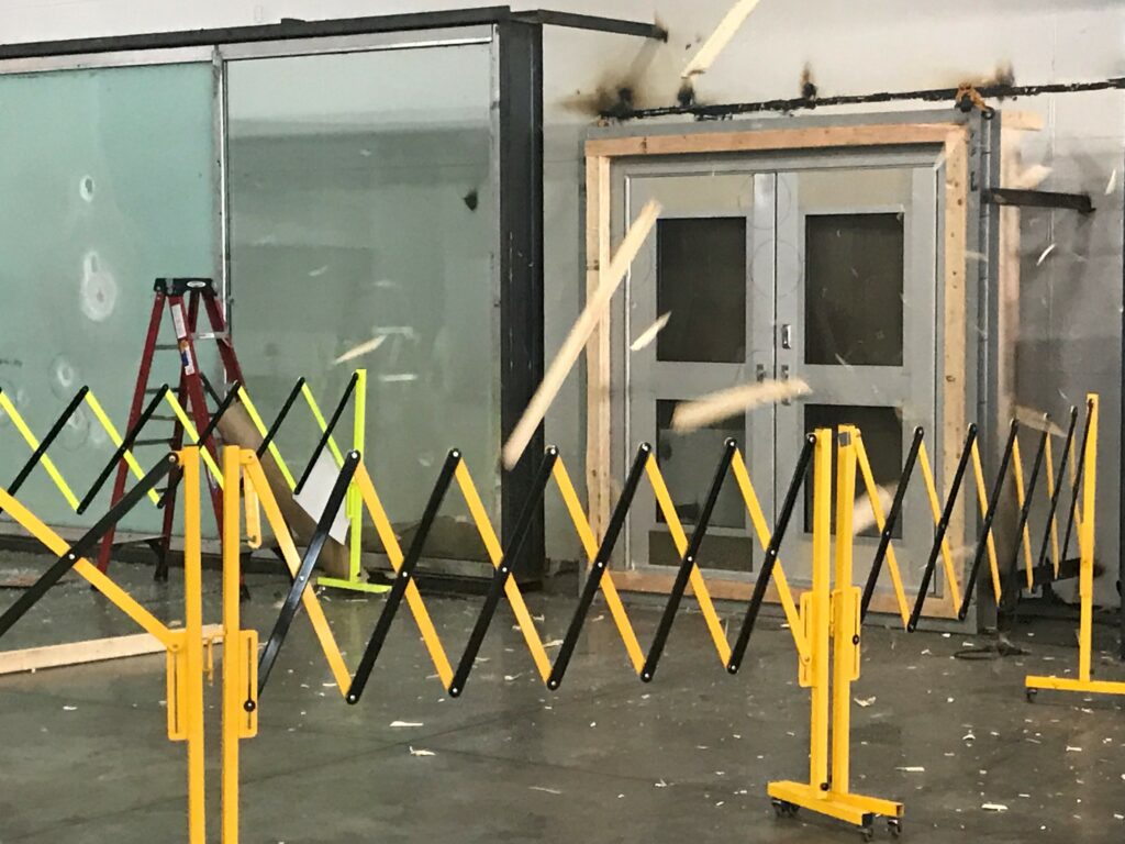 Durante una prueba de simulación, los restos de madera salen disparados hacia una puerta de cristal reforzado. En el entorno industrial se aprecian una barrera amarilla y una escalera roja.